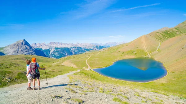 Un par de excursionistas en la cima de la montaña mirando el lago azul y los picos de la montaña. Aventuras de verano en los Alpes. Vista angular amplia desde arriba . —  Fotos de Stock