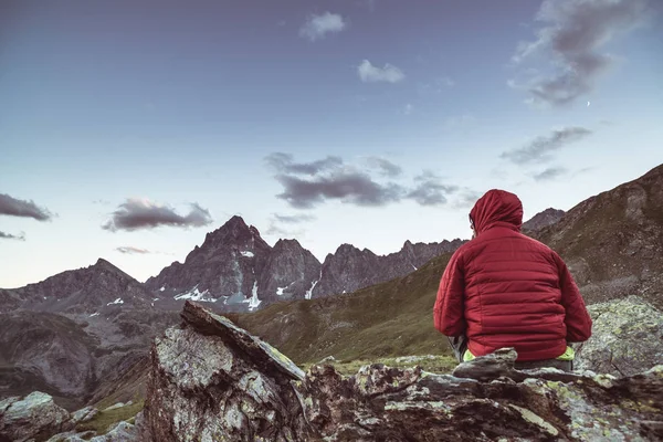 One person sitting on rocky terrain and watching a colorful sunrise high up in the Alps. Wide angle view from above with glowing mountain peaks in the background. Toned image. — Stock Photo, Image