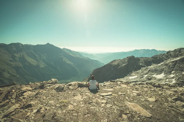 One person sitting on rocky terrain and watching a colorful sunrise high up in the Alps. Wide angle view from above with glowing mountain peaks in the background. Toned image. — Stock Photo, Image