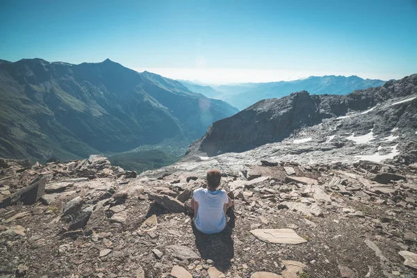 一个人坐在岩石地形上，看着色彩斑斓的朝阳在阿尔卑斯山的高处。宽视角从上面与发光的山峰在背景中。色调的图像. — 图库照片