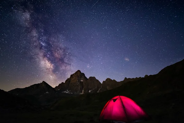 Acampar sob céu estrelado e forma leitosa em alta altitude nos Alpes. Tenda iluminada em primeiro plano e majestoso pico de montanha no fundo. Aventura e exploração no verão . — Fotografia de Stock