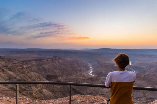 Turista olhando para o Fish River Canyon, destino de viagem cênica no sul da Namíbia. Vista de ângulo ultra grande de cima, pôr do sol cênico colorido . — Fotografia de Stock