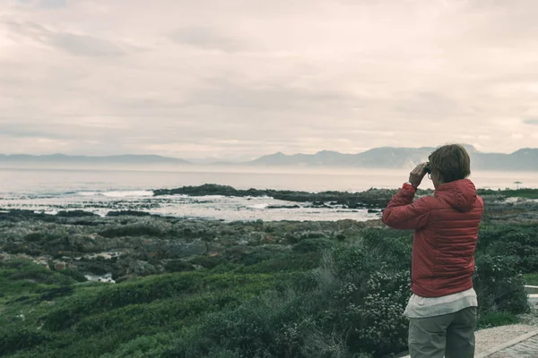 Turisten ser med kikare på den klippiga kusten på De Kelders, Sydafrika, känd för valskådning. Vintersäsongen, molnigt och dramatisk himmel, tonas bild. — Stockfoto