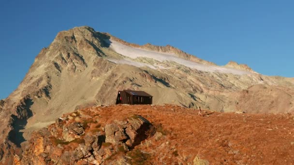 Žena dosáhne horské chatě na vrcholu ve vysoké nadmořské výšce skalnaté krajině s ledovci a peak v pozadí. Dobrodružství na Alpy. — Stock video