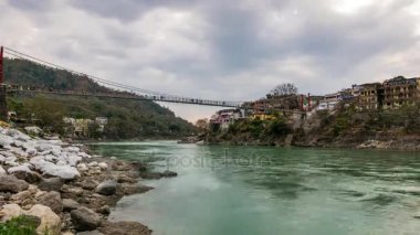 Zaman atlamalı Rishikesh, kutsal şehir, alacakaranlıkta ve hedef Hindistan'da seyahat. Renkli gökyüzü ve Ganj Nehri üzerinde hareketli bulutlar. İnsanlar üzerinde asma köprü geçiş. 