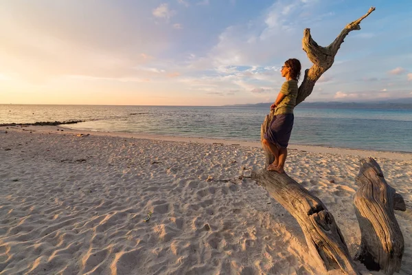 Mulher descansando na árvore esquelética e assistindo a um pôr do sol colorido romântico na praia de Tanjun Karang, Central Sulawesi, Indonésia. Tiro de ângulo largo, exposição longa, movimento turvo . — Fotografia de Stock
