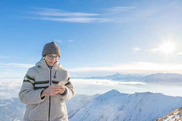 Woman using smart phone on the mountains. Panoramic view of snowcapped Alps in cold winter season. Concept of sharing life moments using new technology.