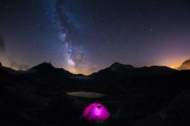 Yıldızlı gökyüzü ve Samanyolu altında Alpler üzerinde yüksek irtifada kamp. Ön plan ve görkemli dağ tepe ve arka planda ışıklı çadırda. Macera ve keşif içinde yaz.