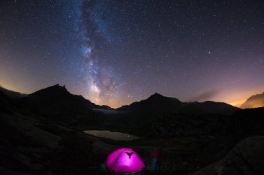 Yıldızlı gökyüzü ve Samanyolu altında Alpler üzerinde yüksek irtifada kamp. Ön plan ve görkemli dağ tepe ve arka planda ışıklı çadırda. Macera ve keşif içinde yaz.