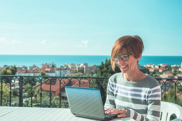 Kvinna med glasögon och casual kläder arbetar på laptop utomhus på terrassen. Vacker bakgrund av gröna kullar och blå himmel i en ljus solig morgon. Tonad bild, riktiga människor. — Stockfoto