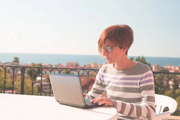 Kvinna med glasögon och casual kläder arbetar på laptop utomhus på terrassen. Vacker bakgrund av gröna kullar och blå himmel i en ljus solig morgon. Tonad bild, riktiga människor. — Stockfoto