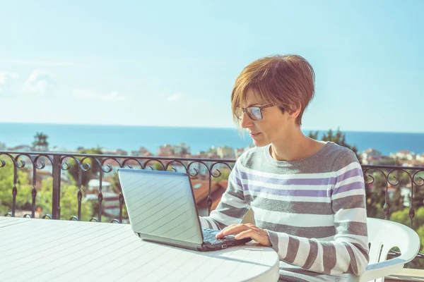 Kvinna med glasögon och casual kläder arbetar på laptop utomhus på terrassen. Vacker bakgrund av gröna kullar och blå himmel i en ljus solig morgon. Tonad bild, riktiga människor. — Stockfoto