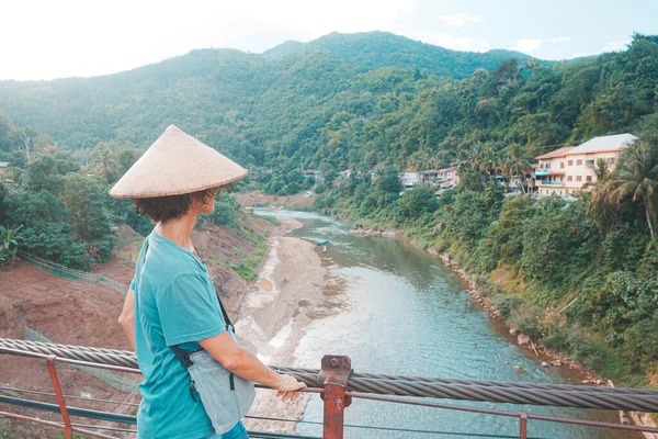 Mujer con sombrero tradicional mirando a la vista en la pasarela obsoleta sobre el río en la aldea de Muang Khua en el norte de Laos, destino de viaje en el sudeste asiático. Tonificado verde azulado estilo naranja —  Fotos de Stock