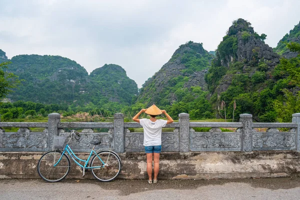 Mujer con sombrero tradicional mirando una vista única de Trang An Ninh Binh, Vietnam. Señora caucásica divirtiéndose en bicicleta de vacaciones en paisaje escénico entre pináculos rocosos y valles verdes . — Foto de Stock