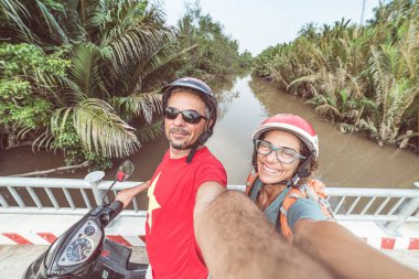 Motosikletle selfie çeken bir çift. Güney Vietnam 'da Mekong Delta bölgesinde kask takan bir adam ve kadın. Yeşilli yeşil hindistan cevizi palmiyesi ağaçları ve su kanalı. Ayarlanmış.