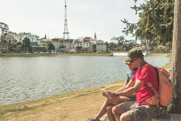 Pareja que utiliza el teléfono inteligente tableta en verde lago parque de la ciudad en la ciudad de Da Lat, Vietnam. Hombre con mochila y mujer con sombrero vietnamita divirtiéndose viajando juntos de vacaciones . — Foto de Stock