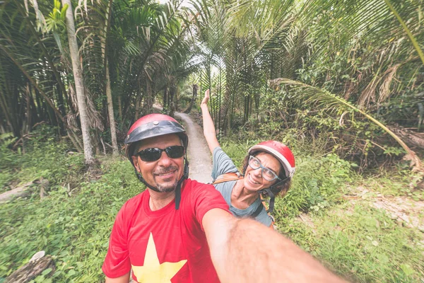 Pareja tomando selfie en moto. Hombre y mujer con casco en bicicleta en la región del delta del Mekong, Vietnam del Sur. Exuberante bosque de palmeras de coco verde y canales de agua. Tonificado . —  Fotos de Stock