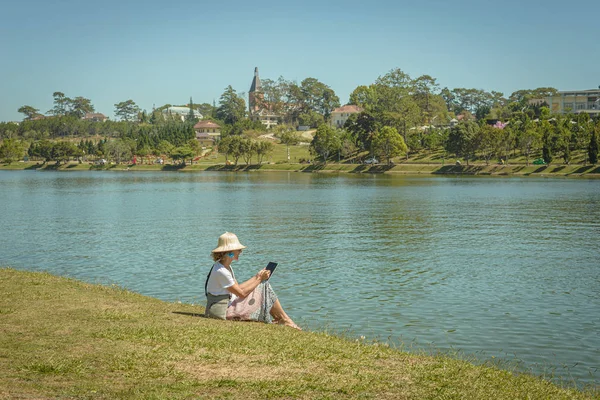 Mujer usando la tableta de teléfono inteligente en verde lago parque de la ciudad en Da Lat ciudad, Vietnam. Señora con sombrero vietnamita divirtiéndose viajando de vacaciones. Tonificado estilo vintage —  Fotos de Stock