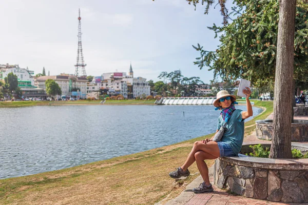 Mujer usando la tableta de teléfono inteligente en verde lago parque de la ciudad en Da Lat ciudad, Vietnam. Señora con sombrero vietnamita tomando selfie divertirse viajando de vacaciones . — Foto de Stock