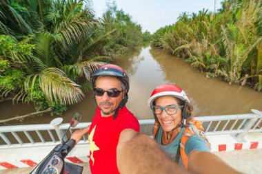 Motosikletle selfie çeken bir çift. Güney Vietnam 'da Mekong Delta bölgesinde kask takan bir adam ve kadın. yemyeşil hindistan cevizi palmiyesi ağaçları ve su kanalları. Ayarlanmış.