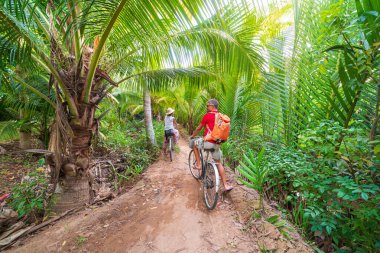 Mekong Delta bölgesinde bisiklet süren turist çift, Ben Tre, Güney Vietnam. Kadın ve erkek, yeşil tropikal ormanlık ve hindistan cevizi palmiyeleri arasında bisiklet sürerken eğleniyorlar. Arka görünüm.