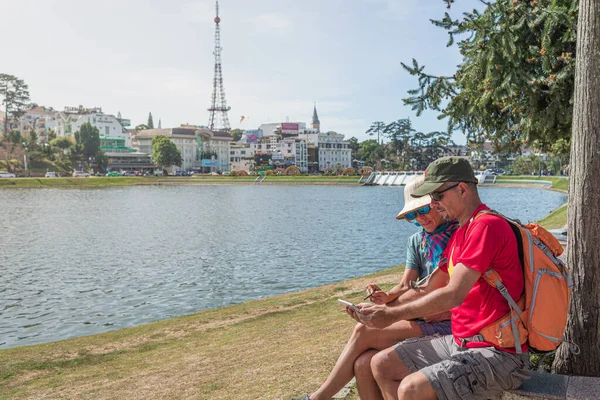 ベトナムのダラット町の緑の都市公園湖でタブレットスマートフォンを使用してカップル バックパックを持つ男とベトナムの帽子を持つ女性楽しい旅行を一緒に休暇 — ストック写真