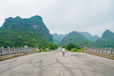 Bisikletle gezen bir kadın manzaralı bir yolda, kayalıkların ve yeşil vadilerin arasında tek başına. Trang An Ninh Binh 'in eşsiz manzarası, Vietnam. Sosyal uzaklık, yalnız seyahat, alternatif hareket.