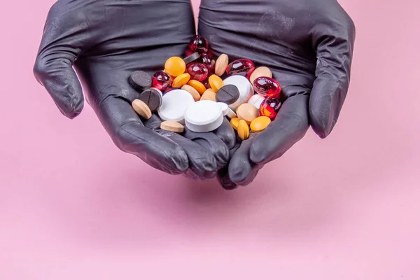Manos de hombre en guantes de goma negro sostienen muchas píldoras multicolores sobre un fondo rosa —  Fotos de Stock