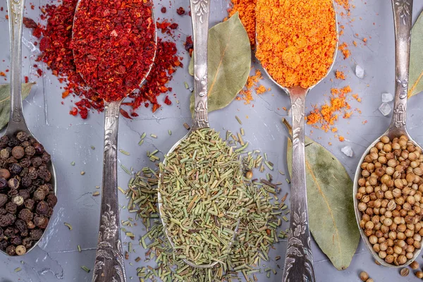 Turmeric powder, paprika, coriander and black peppercorns in metal teaspoons on a gray background