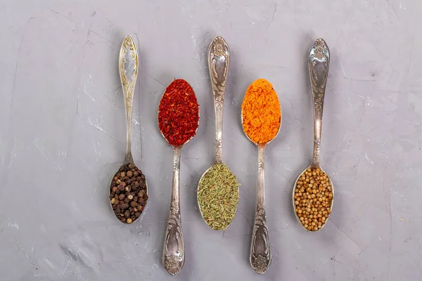 Turmeric powder, paprika, coriander and black peppercorns in metal teaspoons on a gray background