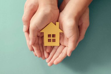 Hands holding yellow paper house on blue background, family home clipart