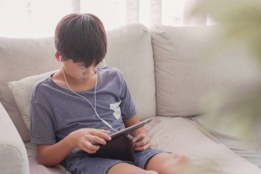 Genç Asyalı çocuk evde dijital tablet kullanıyor, podcast, oyun, online eğitim, elearning, ev eğitimi, sosyal uzaklık, izolasyon, tecrit kavramı dinliyor.