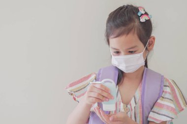 Maske takan ve el dezenfektanı kullanan Asyalı bir kız, okul yeniden açılıyor, covid-19 koronavirüs salgını bittikten sonra okula geri dönüyor, yeni normal konsept.