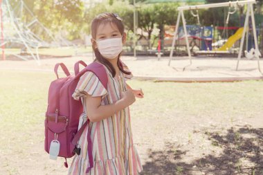 Maske takan ve okul çantasıyla oyun parkının yanında el dezenfektanı taşıyan karma Asyalı kız okul açılışı, covid-19 Coronavirus salgını bittikten sonra okula dönmek, yeni normal konsept.