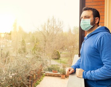 Coronavirus covid19 salgınından dolayı karantina sırasında maskeli genç beyaz adam terasa bakıyor..