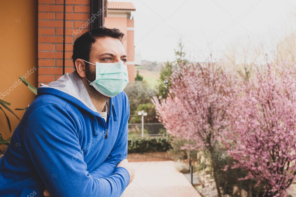 Young caucasian man with mask looking out onto home terrace during quarantine due to coronavirus covid19 pandemic.