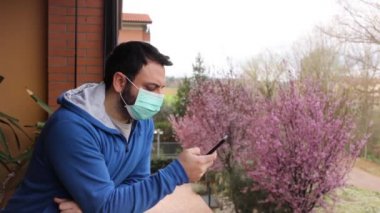 Koronavirüs salgını yüzünden karantina sırasında cep telefonu kullanarak terasa bakan maskeli beyaz bir genç..