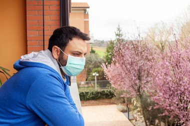 Coronavirus covid19 salgınından dolayı karantina sırasında maskeli genç beyaz adam terasa bakıyor..