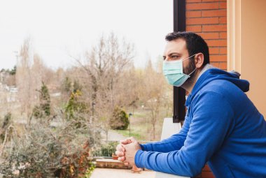 Coronavirus covid19 salgınından dolayı karantina sırasında maskeli genç beyaz adam terasa bakıyor..