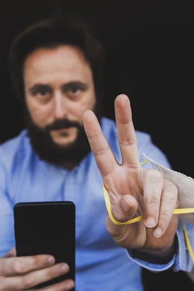 End of quarantine. All the bad times will come to an end, and the good ones will be back soon. Bearded man holding a smartphone does victory sign; he is hanging a medical mak on his hand. Selective focus, dark background
