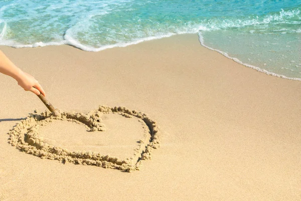 Hand of woman drawing heart on sand seashore near wavy sea. Vacation concept. Copy space on right side