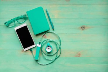 Portatif akü, çalar saat, kağıt defter, tahta masa üzerinde kalem ve gözlük ile şarj edilmiş akıllı telefon, hepsi turkuaz renkli. Güç bankasından şarj edilmiş telefon. Rutin yaşam tarzı kavramı.