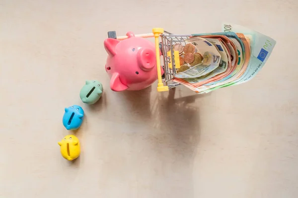 Piglet banks following big pig bank, which rolling shopping cart with euro notes on wood background. Big pig, little pig banks and euro banknotes in shopping trolley. Top view. Saving money concept