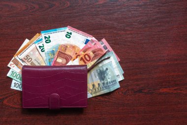 Ahşap arka planda euro banknotları olan deri cüzdan. Masada deri cüzdanı olan çeşitli avroluk banknotlar. Mor cüzdan ve maun masa üzerinde euro banknotları. Cüzdan ve maaş. Nakit ödeme kavramı