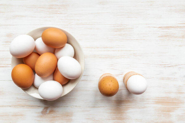 Bowl with brown and white chicken eggs and boiled eggs in holders on table. Eggs in plate and holders on wood background. Free-range organic eggs. Top view, copy space. Natural healthy food concept.