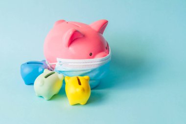 Pembe domuz kumbarası, koruyucu yüz maskesi takıyor ve küçük kumbaraların etrafında, mavi arka planda izole edilmiş. Tıbbi maskeli domuzcuk kumbarası ve diğerleri. Koronavirüs salgını sırasında para tasarrufu kavramı.