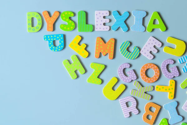 Word Dyslexia written of wooden letters and scattered letters below on azure background. Inscription DYSLEXIA formed with colorful wooden letters on blue backdrop. Reading difficulties concept.