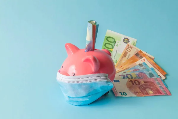 Pembe domuz kumbarası, koruyucu maske takıyor ve mavi arka planda izole edilmiş euro banknotları. Tek kullanımlık tıbbi maskeli ve euro banknotlu domuz kumbarası. Koronavirüs salgını sırasında para tasarrufu kavramı.