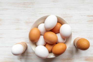 Tabakta kahverengi ve beyaz yumurtalar ve ahşap arka planda tutucular. Tahta masanın üzerinde kahverengi ve beyaz tavuk yumurtaları olan bir kase. Serbest organik yumurtalar. Düz konum, kopyalama alanı. Doğal sağlıklı gıda konsepti.