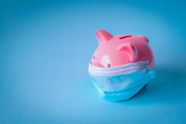 Pembe domuz kumbarası, koruyucu yüz maskesi takıyor, mavi arka planda izole edilmiş. Tek kullanımlık tıbbi maskeli domuz kumbarası. Koronavirüs salgını sırasında para tasarrufu kavramı.
